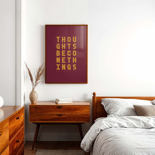 Minimal burgundy and mustard typography print with the words “Thoughts become things, make them brilliant” displayed in bold type on a white bedroom wall.