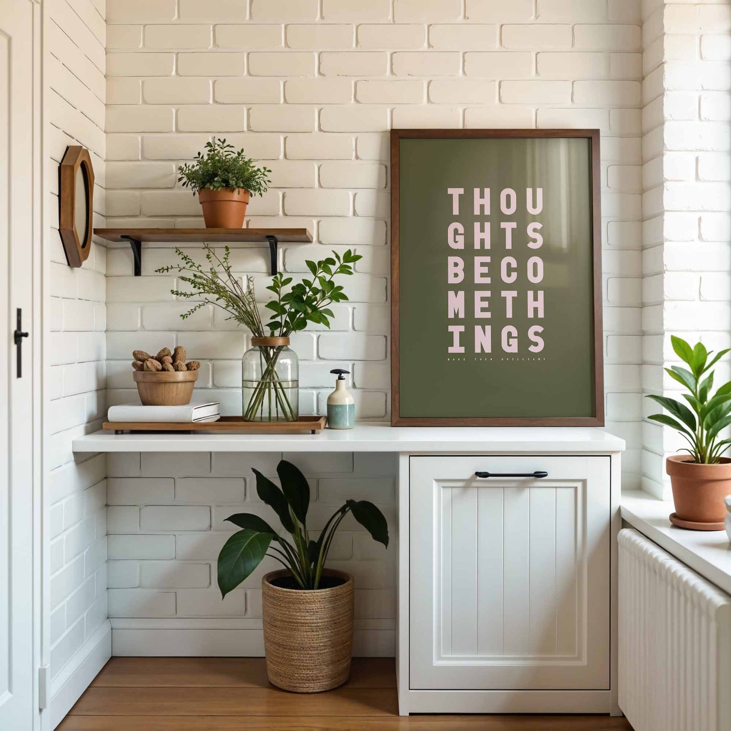 Minimal olive and pink typography print with the words “Thoughts become things, make them brilliant” displayed in bold type on a neutral kitchen brick wall.