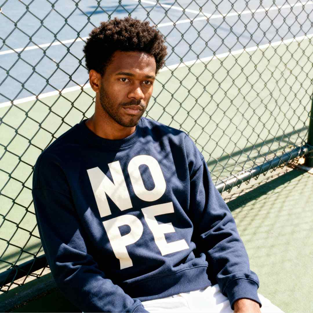 Black man leaning up against a fence wearing a navy NOPE sweatshirt

