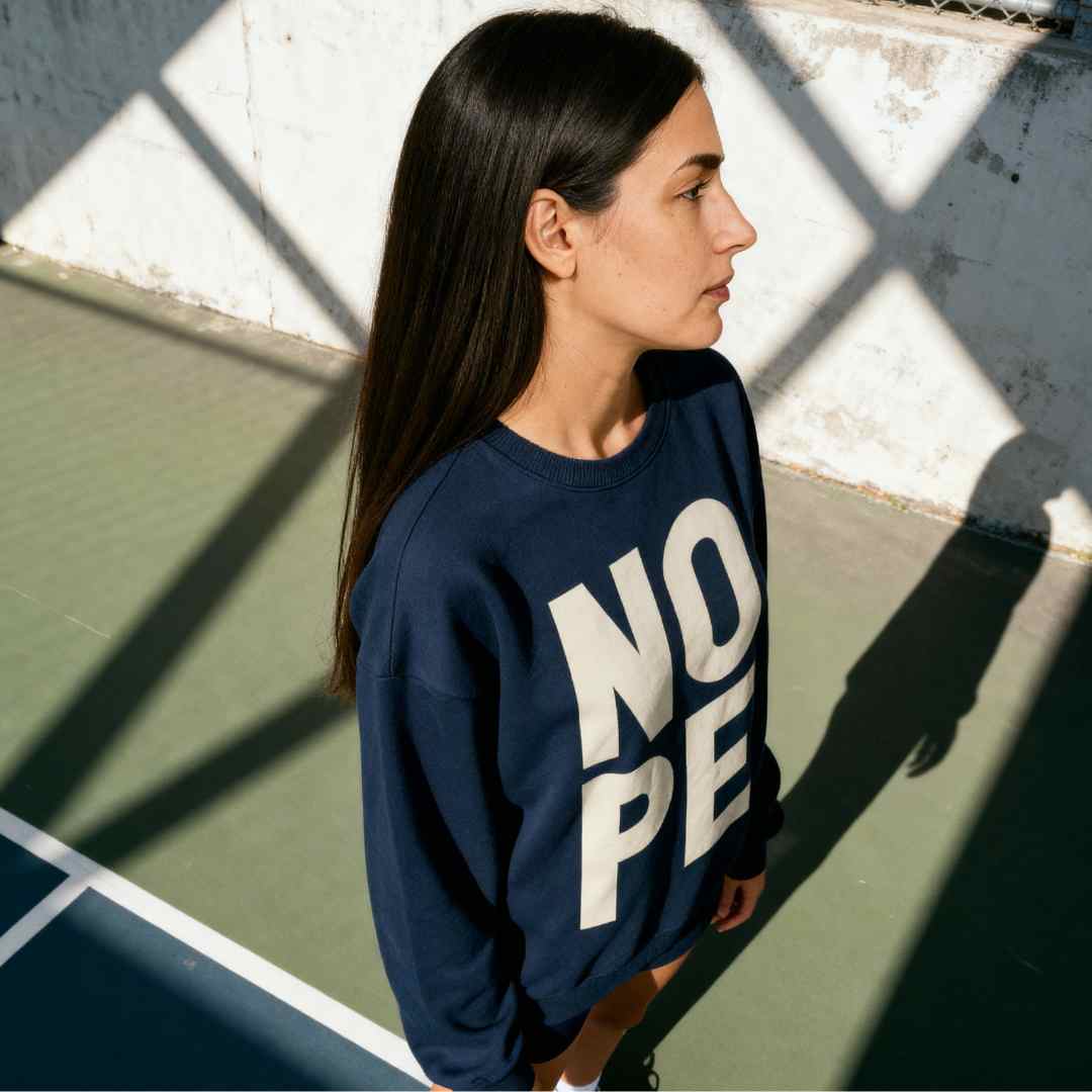 Olive skinned woman with dark long hair wearing oversized NOPE sweatshirt in Navy, styled on a vintage tennis court

