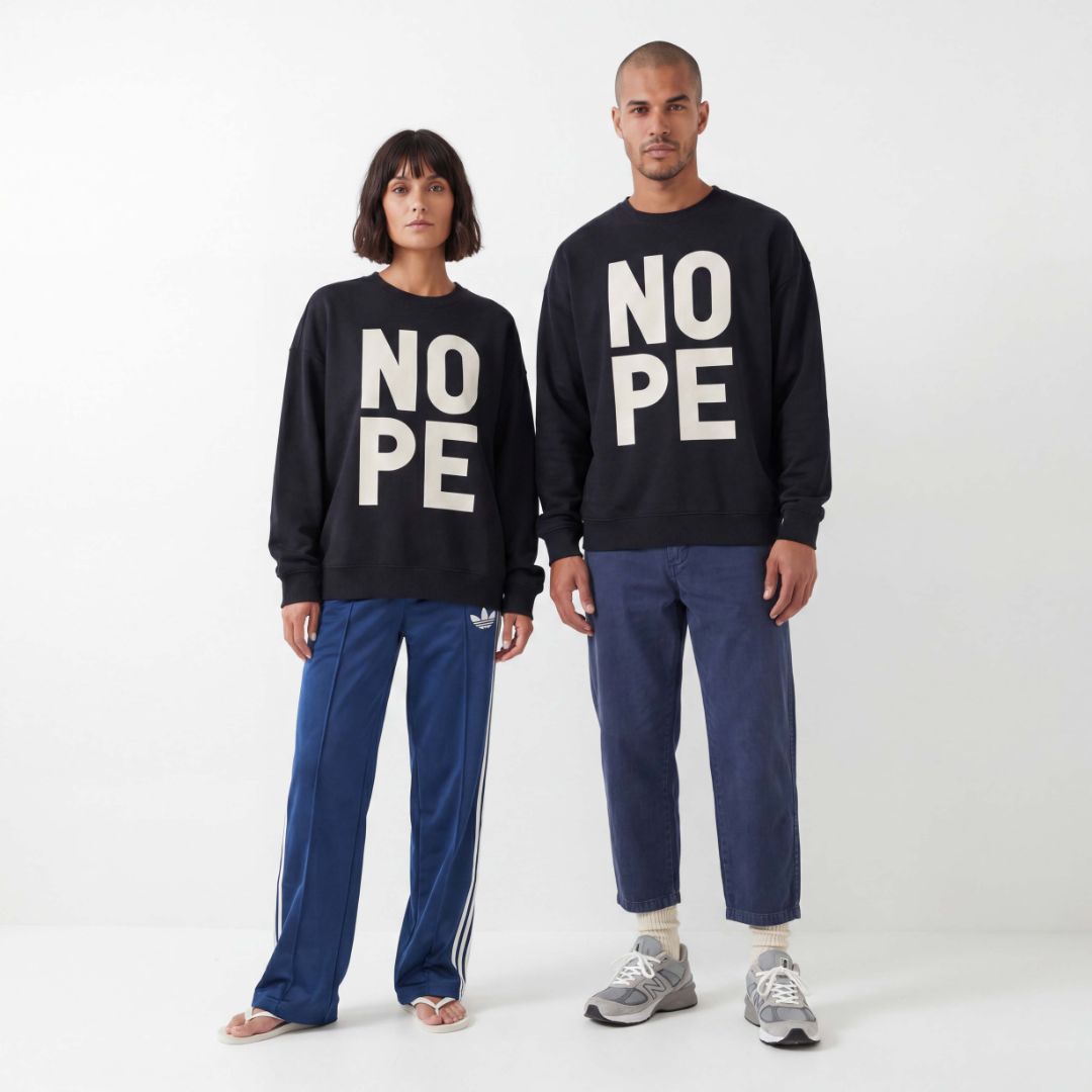 Man and woman wearing unisex sweatshirt with large letters saying NOPE, shot in a minimal studio. Relaxed outfit styling with adidas track pants and barel leg jeans. 
