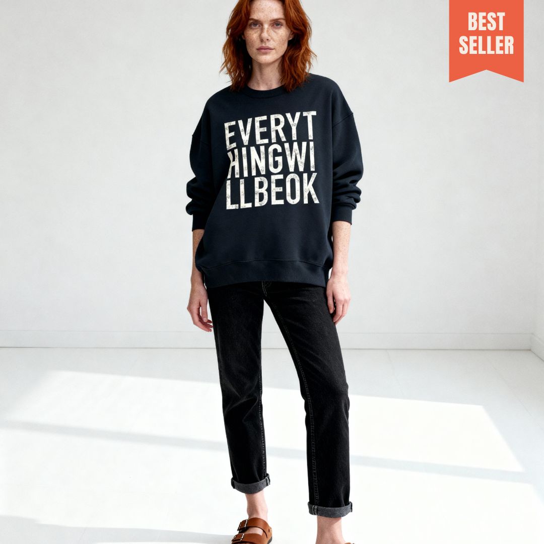 Auburn haired model with freckles, in minimal studio wearing oversized black sweatshirt with bold white typographic design "Everything will be ok"