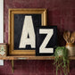 Oil painted bold typography letter initial prints A & Z styled on a kitchen shelf against burgundy tiles and vintage utencils. 