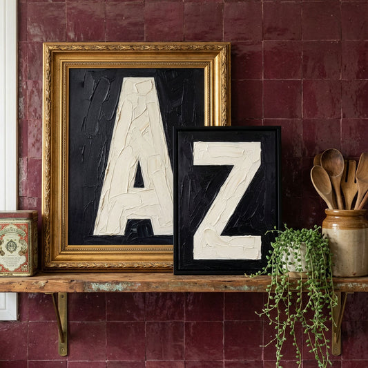 Oil painted bold typography letter initial prints A & Z styled on a kitchen shelf against burgundy tiles and vintage utencils. 