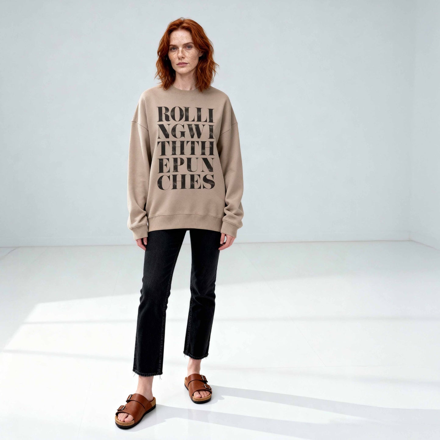 Driftwood coloured oversized sweatshirt with Rolling With The Punches typography design styled with jeans on auburn haired model in minimalist studio. 