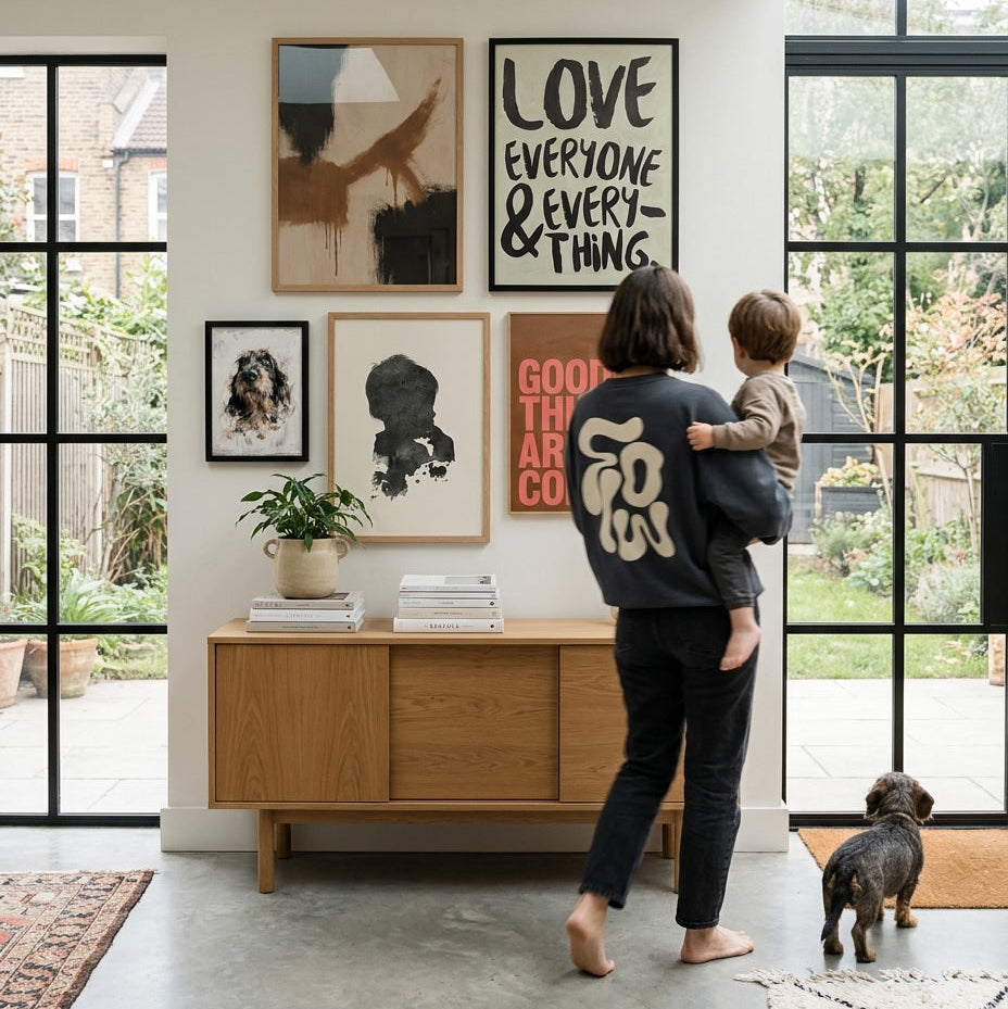 A woman carrying a child walks through a bright family home featuring a gallery wall of personalised portrait artwork, typography prints and framed illustrations above a wooden sideboard, showcasing Burnt Peach Studio's thoughtfully designed wall art.