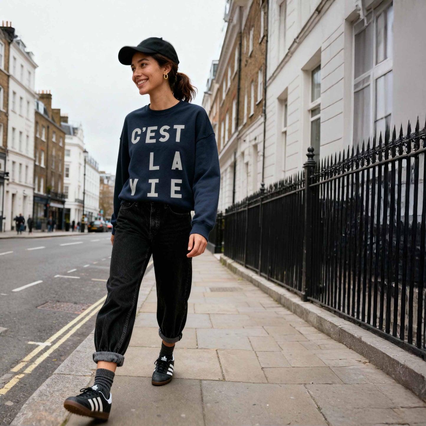 Brunette modeling navy "C'est La Vie' statement sweatshirt' tucked into jeans walking down a london street.  