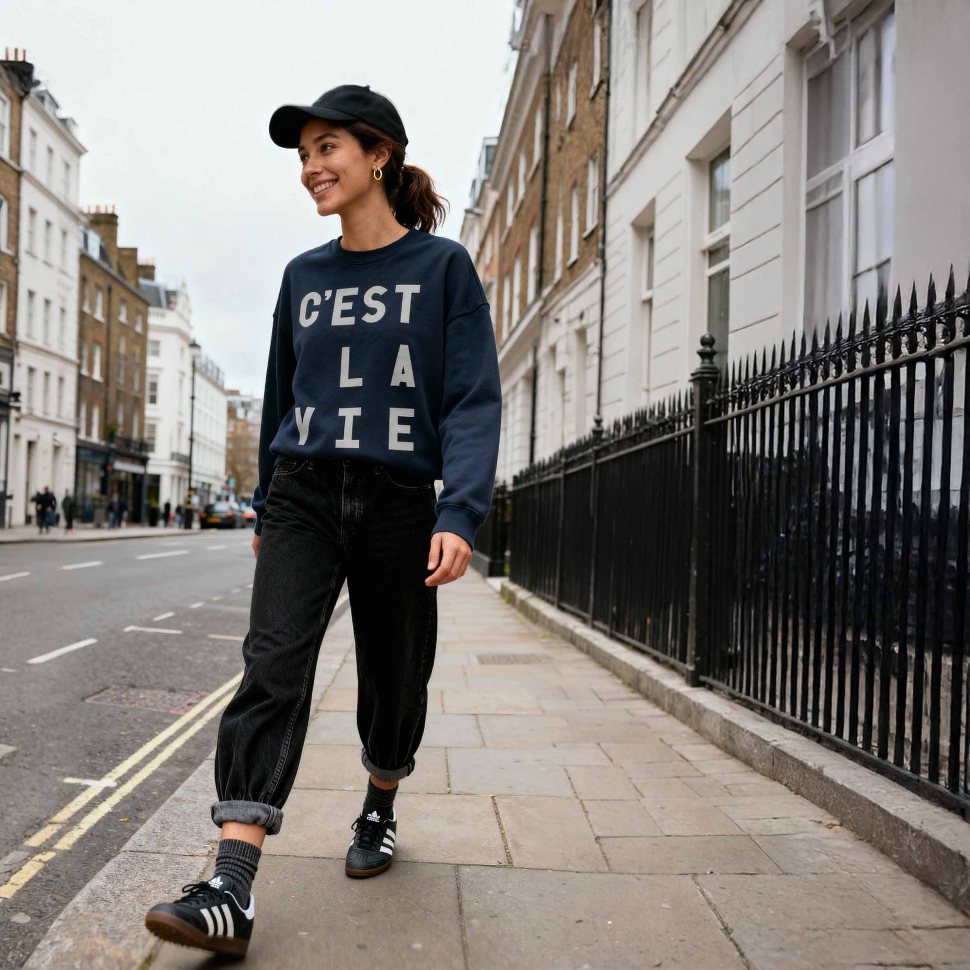 Brunette modeling navy "C'est La Vie' statement sweatshirt' tucked into jeans walking down a london street.  