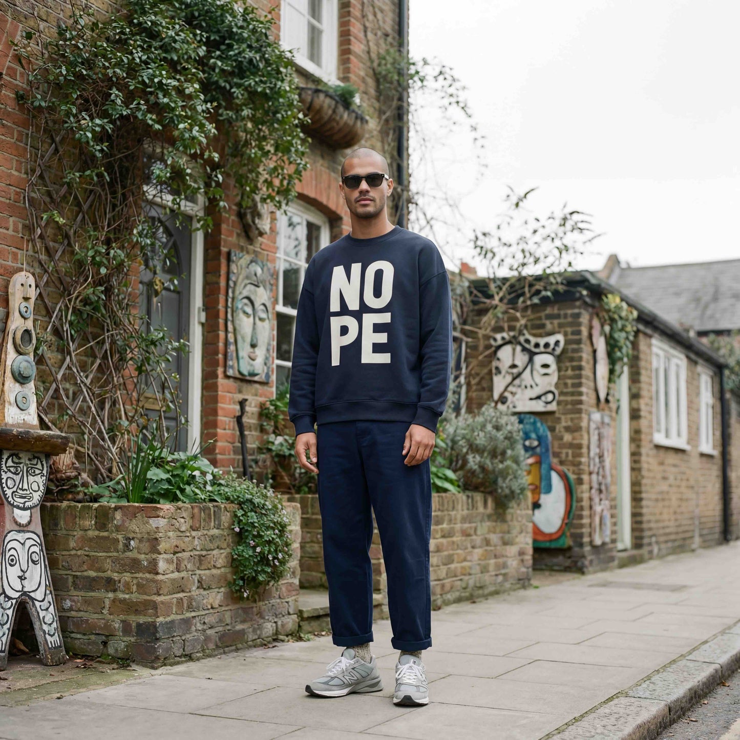 Man wearing NOPE navy sweatshirt standing outside a london home. Styling the sweatshirt with barel leg trousers and new balance trainers and sunglasses. 