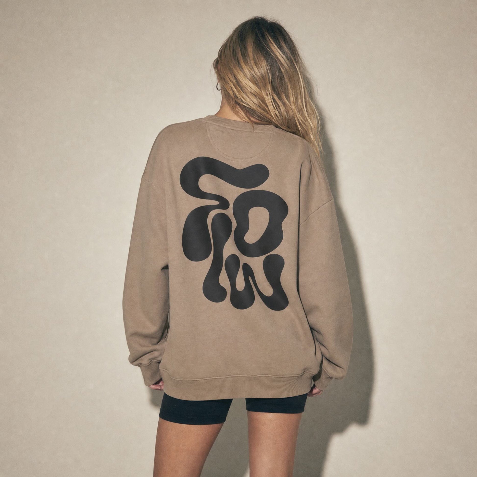 Model wearing Flow sweatshirt by BurntPeach in colour driftwood, bold typographic design on the reverse, shot in a studio. 