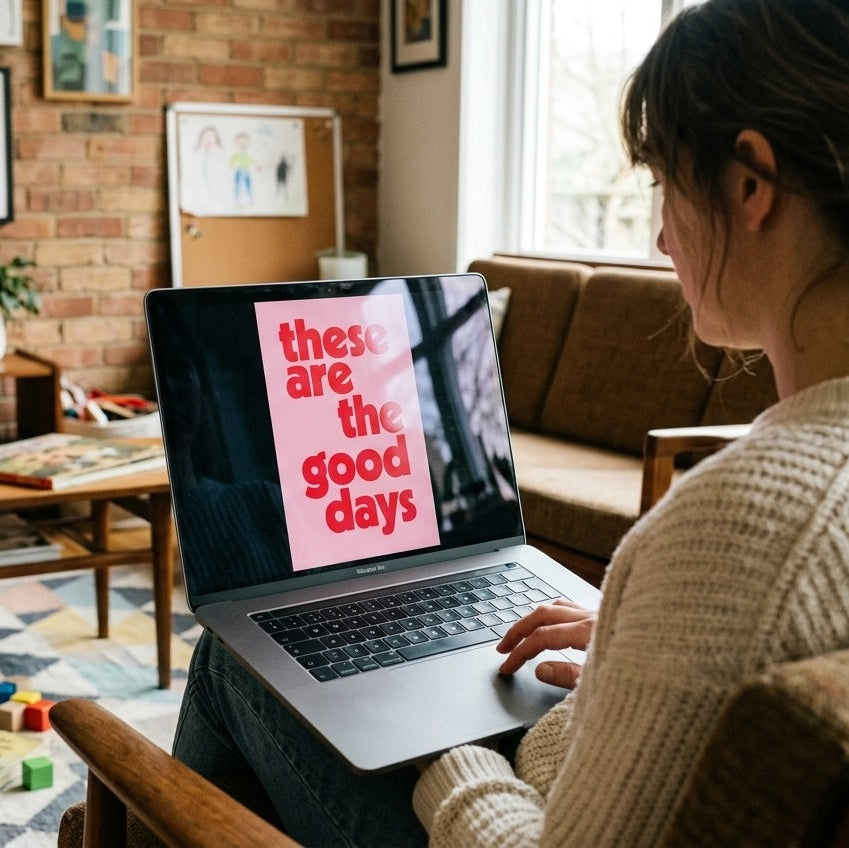 Woman viewing a Burnt Peach digital download on a laptop in a family living room. The "These Are the Good Days" typography print is shown on screen, demonstrating printable wall art for modern gallery walls and home décor.