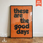 Orange and black “These Are The Good Days” typography art print in a slim black frame, leaning against a white wall on a parquet wooden floor.