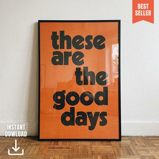 Orange and black “These Are The Good Days” typography art print in a slim black frame, leaning against a white wall on a parquet wooden floor.