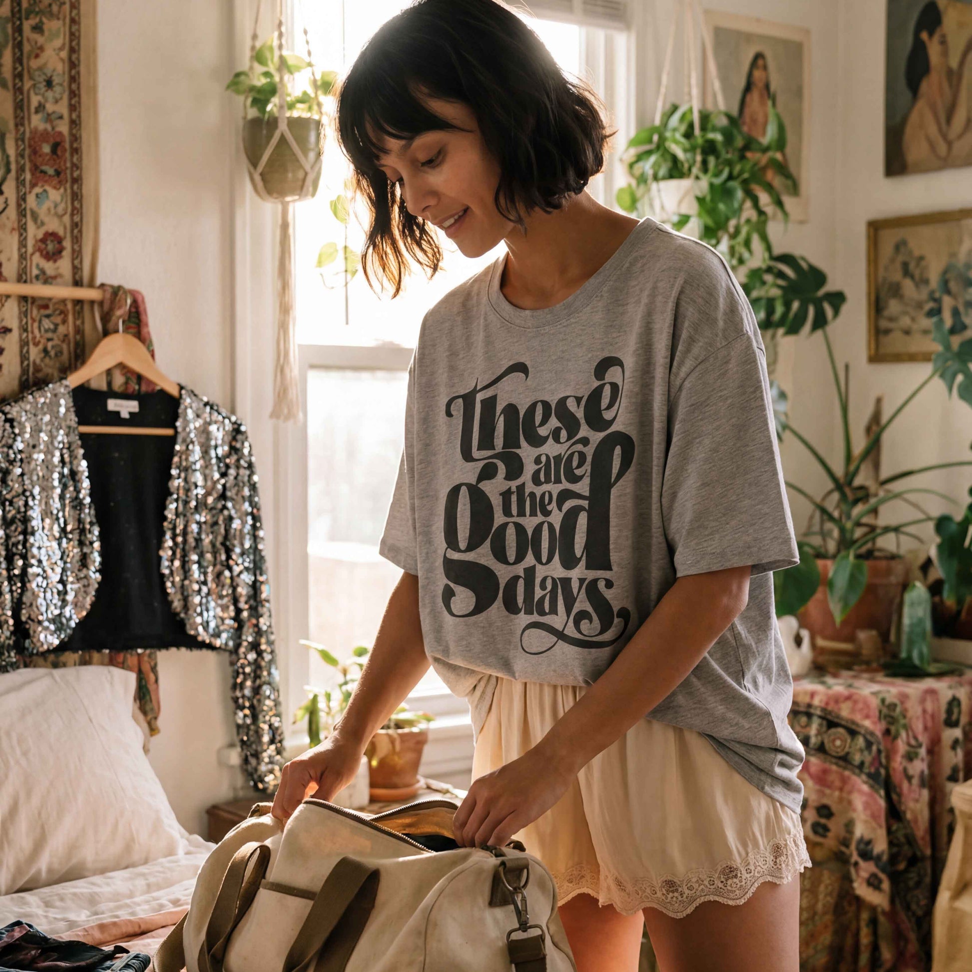 Girl packing her bag in bedroom, wearing oversized grey 'These are the good days" tshirt with silk shorts. 