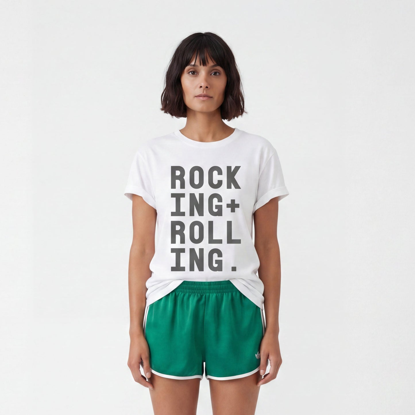 model in white studio wearing a white tshirt with black text Rocking + Rolling styled with green adidas sports shorts. 