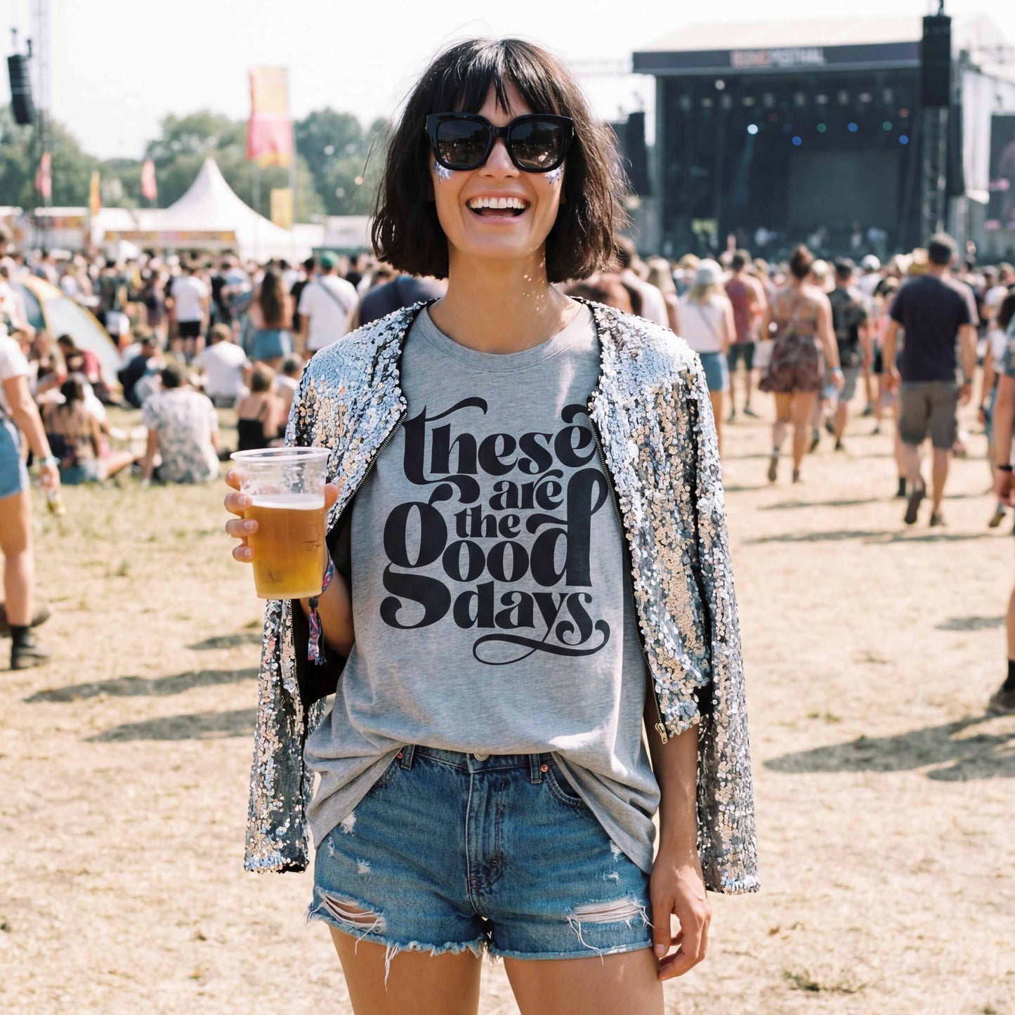 Woman wearing “These Are the Good Days” graphic t-shirt at a summer festival, styled casually with denim shorts and sunglasses