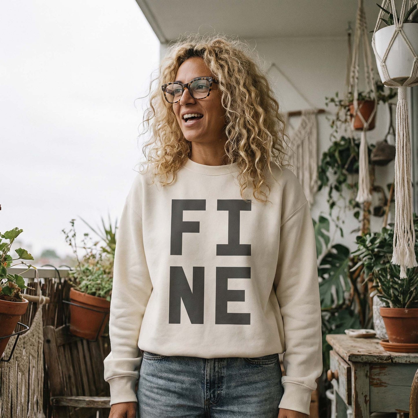 Curly haired woman wearing ivory coloured  bold typographic designed sweatshirt, with 'FINE' in black.Styled on a balcony with plants. 