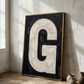 Letter G oil paint art print framed in wood leaning against a textured plaster wall interior with large window