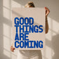 Woman holding up the print Good Things Are Coming with bold blue typography on cream background. 