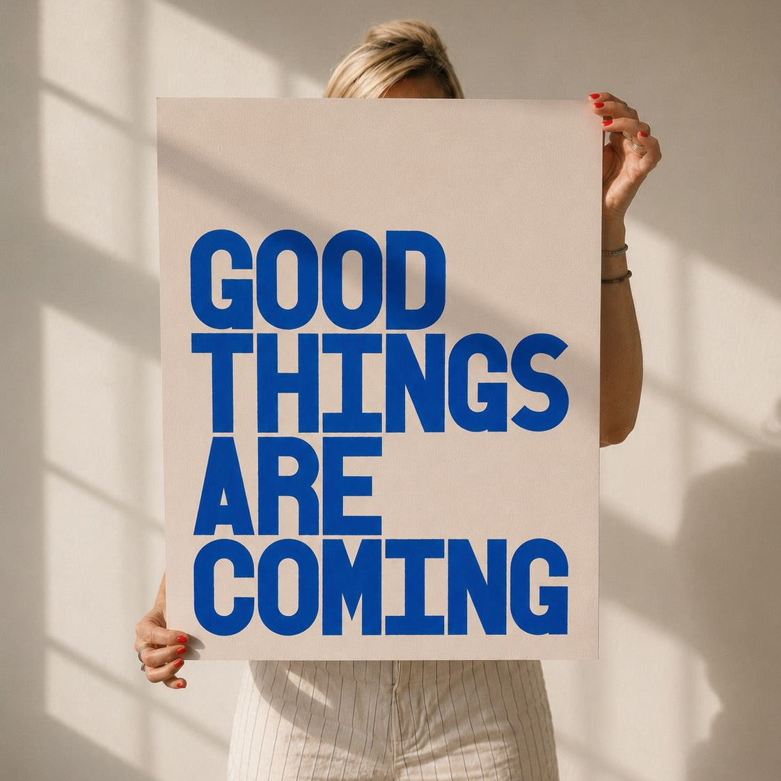 Woman holding up the print Good Things Are Coming with bold blue typography on cream background. 