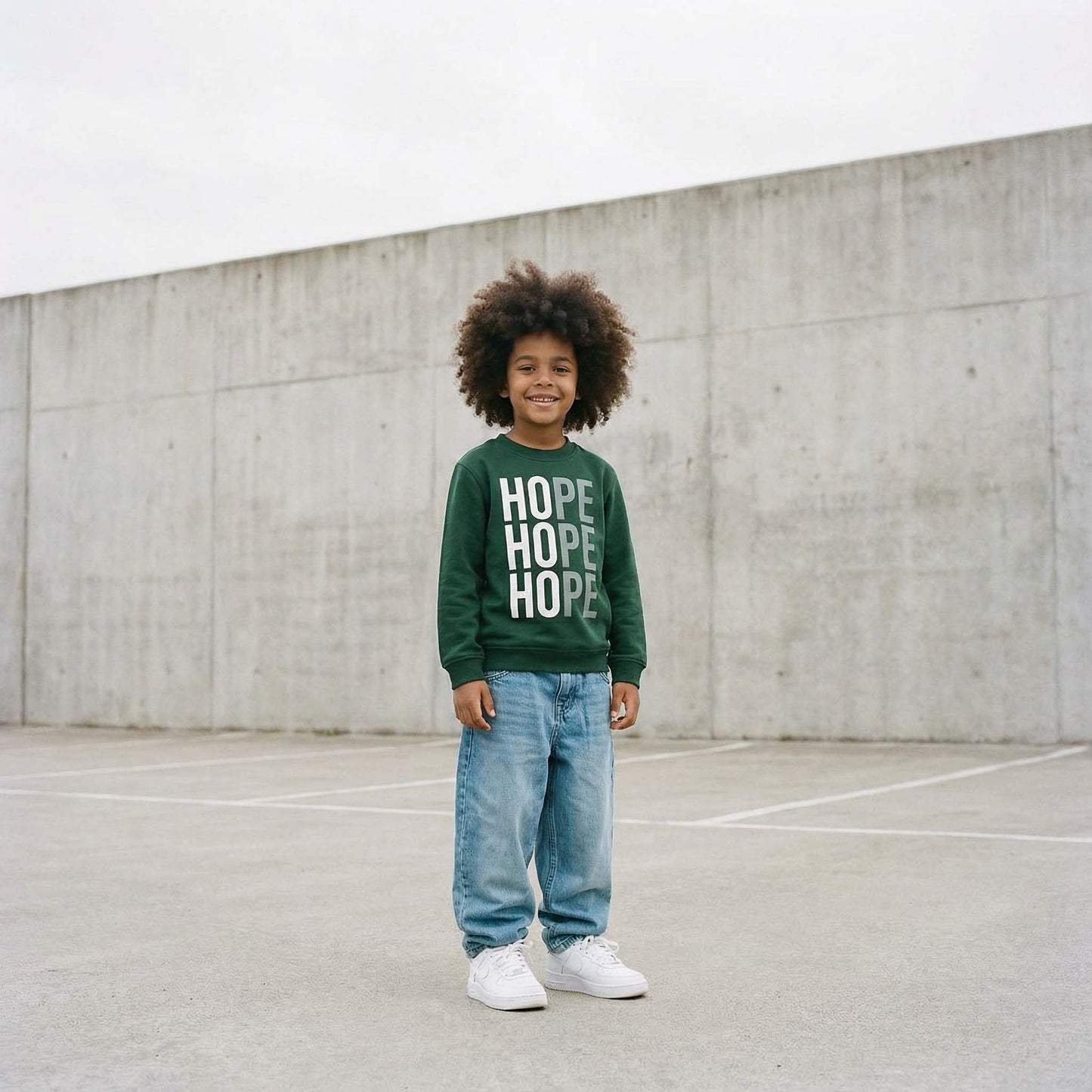 Little boy with big afro hair wearing Hope sweatshirt in bottle green with white design. photographed against a concrete wall.  Sweatshirt raising money for Centrepoint charity. 