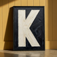 Letter K oil paint art print framed in black styled leaning against a mustard coloured panneled wood wall, with natural shadows