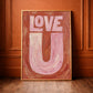 Love U typography created in oil paint, in pinks and orage, framed in brass leaning agaist a orange panel wall. 
