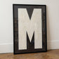 Large textured letter M alphabet print featuring bold cream typography on a black painted background, framed in black.