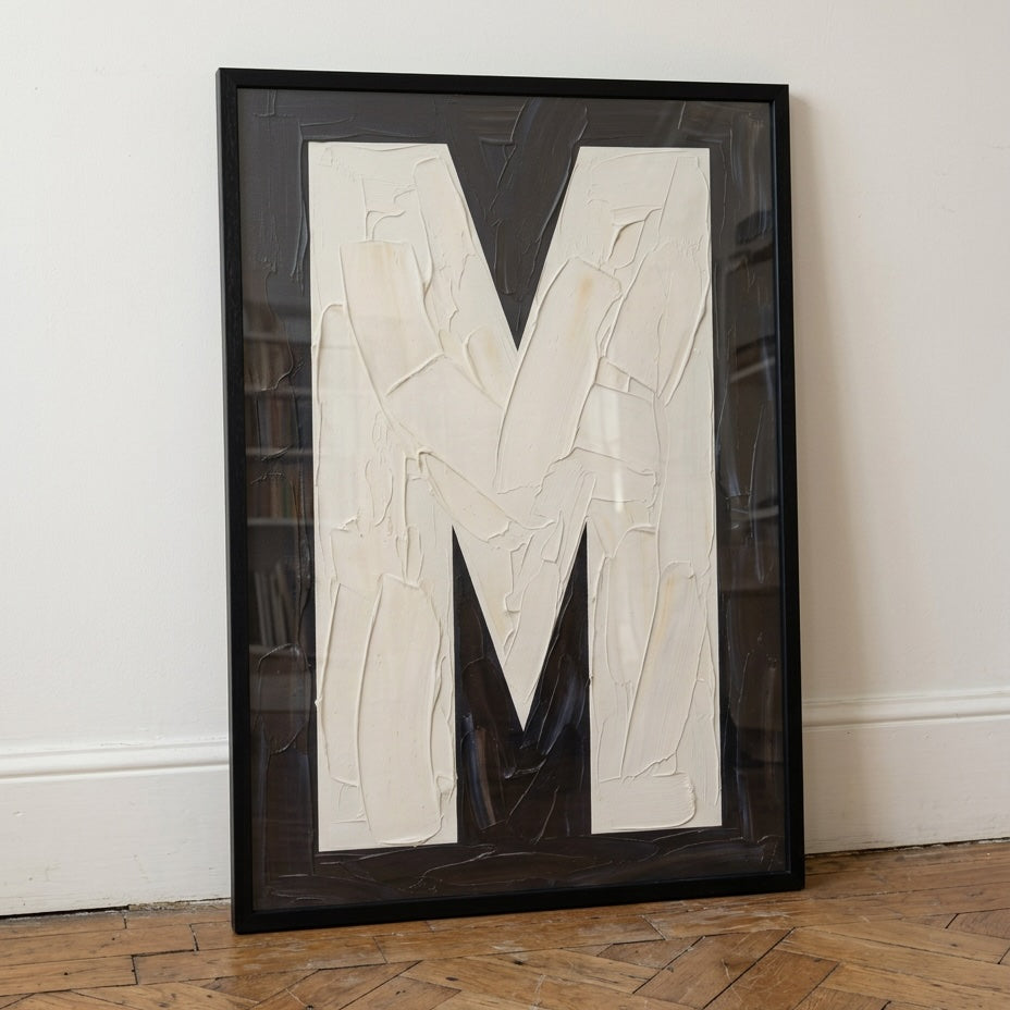Large textured letter M alphabet print featuring bold cream typography on a black painted background, framed in black.