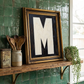 Letter M oil paint art print framed in a vintage gold frame, on a shelf against  green zellige tiled kitchen