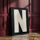 Letter N oil paint art print framed in a wooden frame, leaning against burgundy panneled ornate wall. 