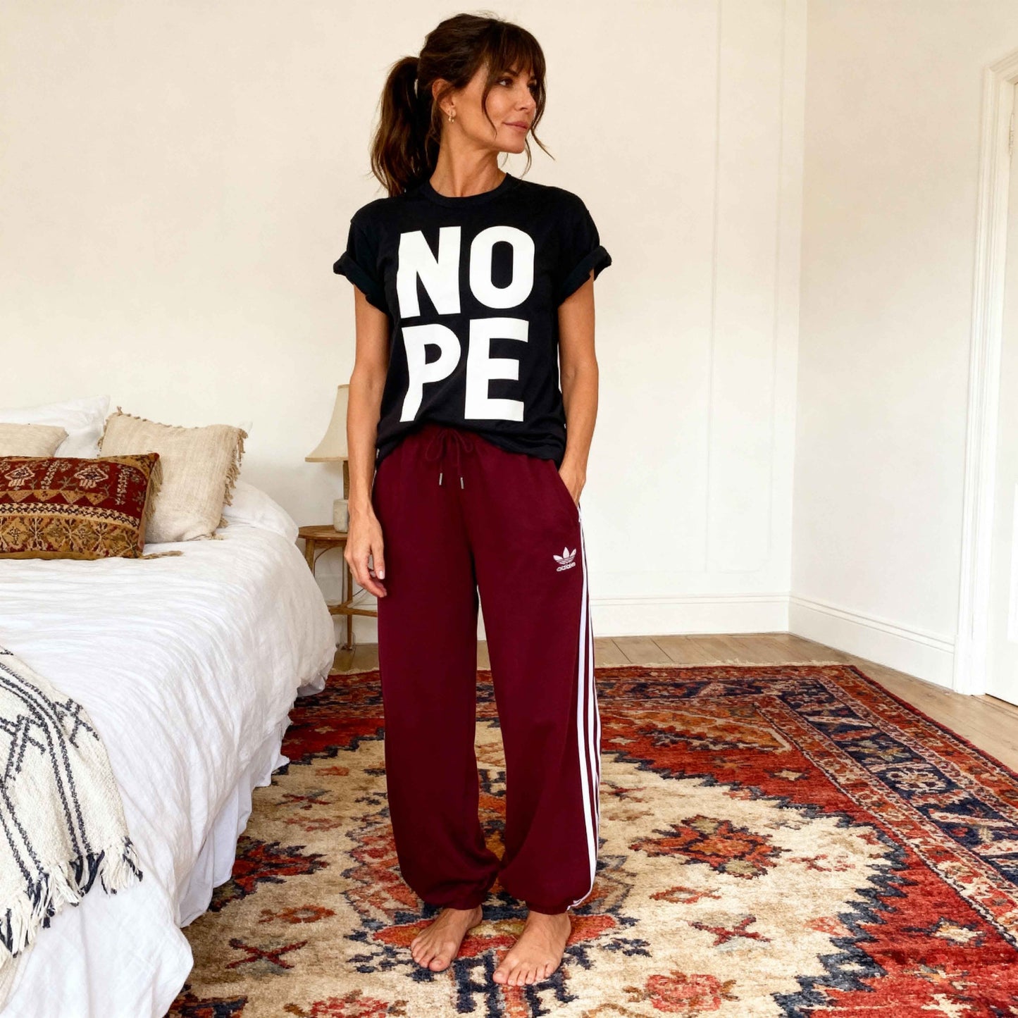 Model in boho bedroom wearing black Nope tshirt with rolled sleeves, and maroon adidas jogging bottoms. 