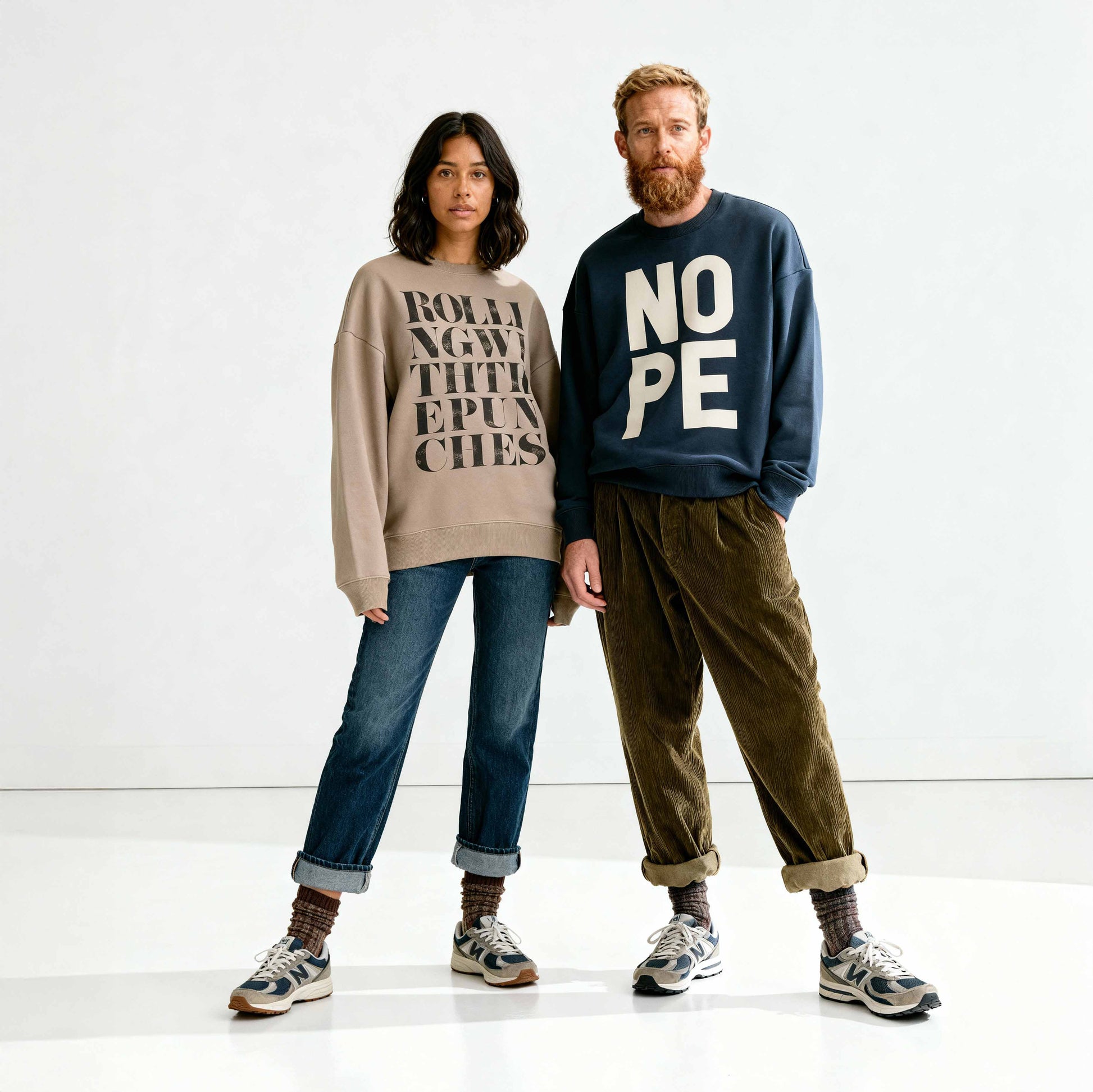 Couple modeling oversized, statement sweatshirts with bold typography designs NOPE and Rolling with the punches, in minimalist studio. 