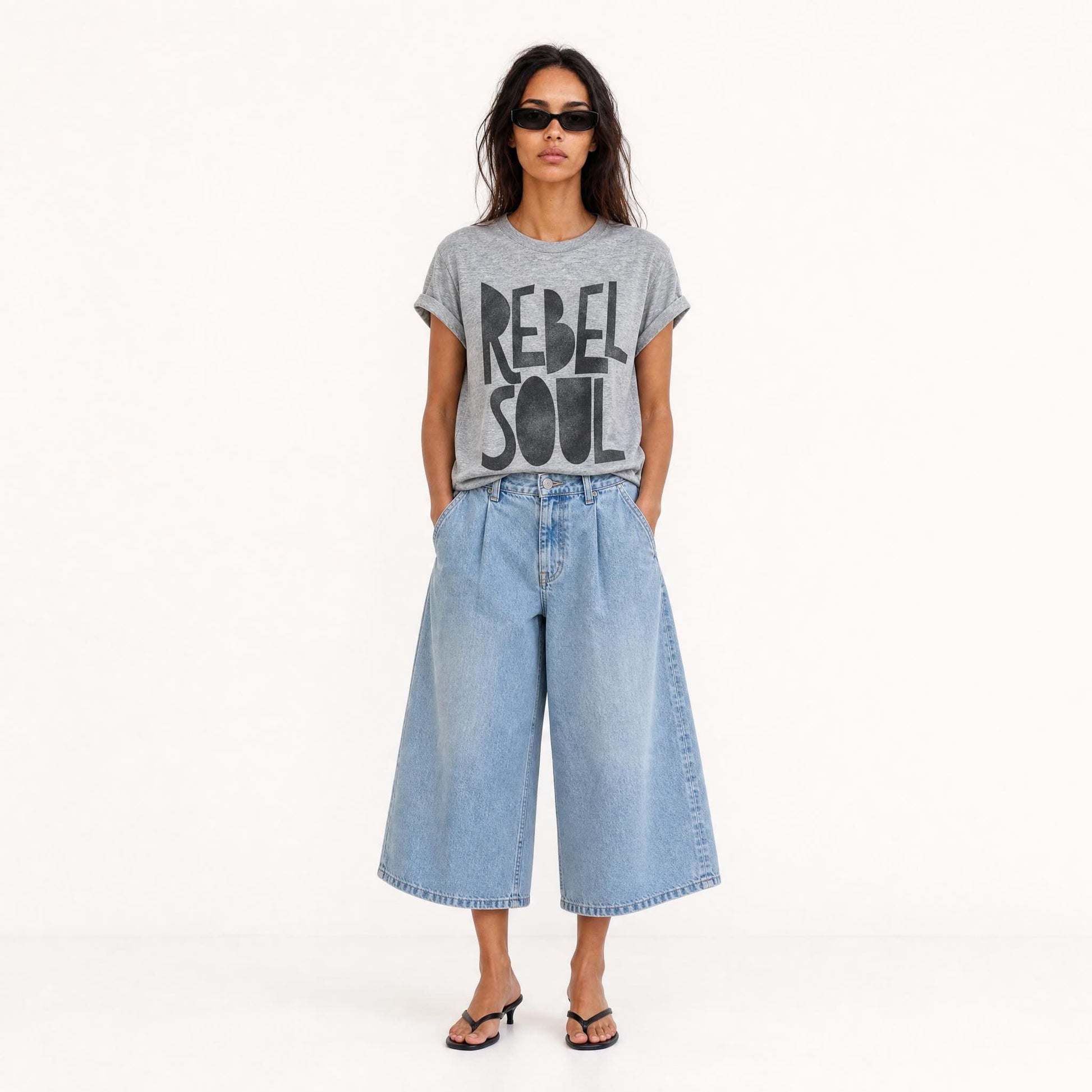 Woman wearing an oversized grey Rebel Soul graphic T-shirt with bold black typography, styled with light blue wide-leg cropped jeans and black kitten heel sandals.