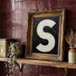 Letter S oil paint art print framed in a vintage ornate gold frame, on a shelf against  burgundy zellige tiled kitchen