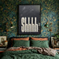 Shhh black textured bedroom wall art print in a black frame above a bed with botanical wallpaper and green bedding.