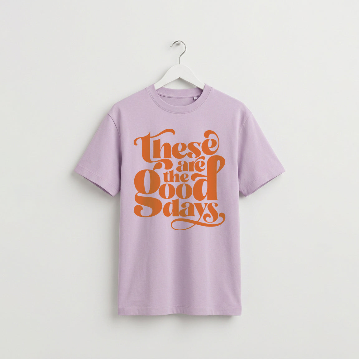Lavender “These Are the Good Days” graphic t-shirt on white wooden hanger, minimalist studio background