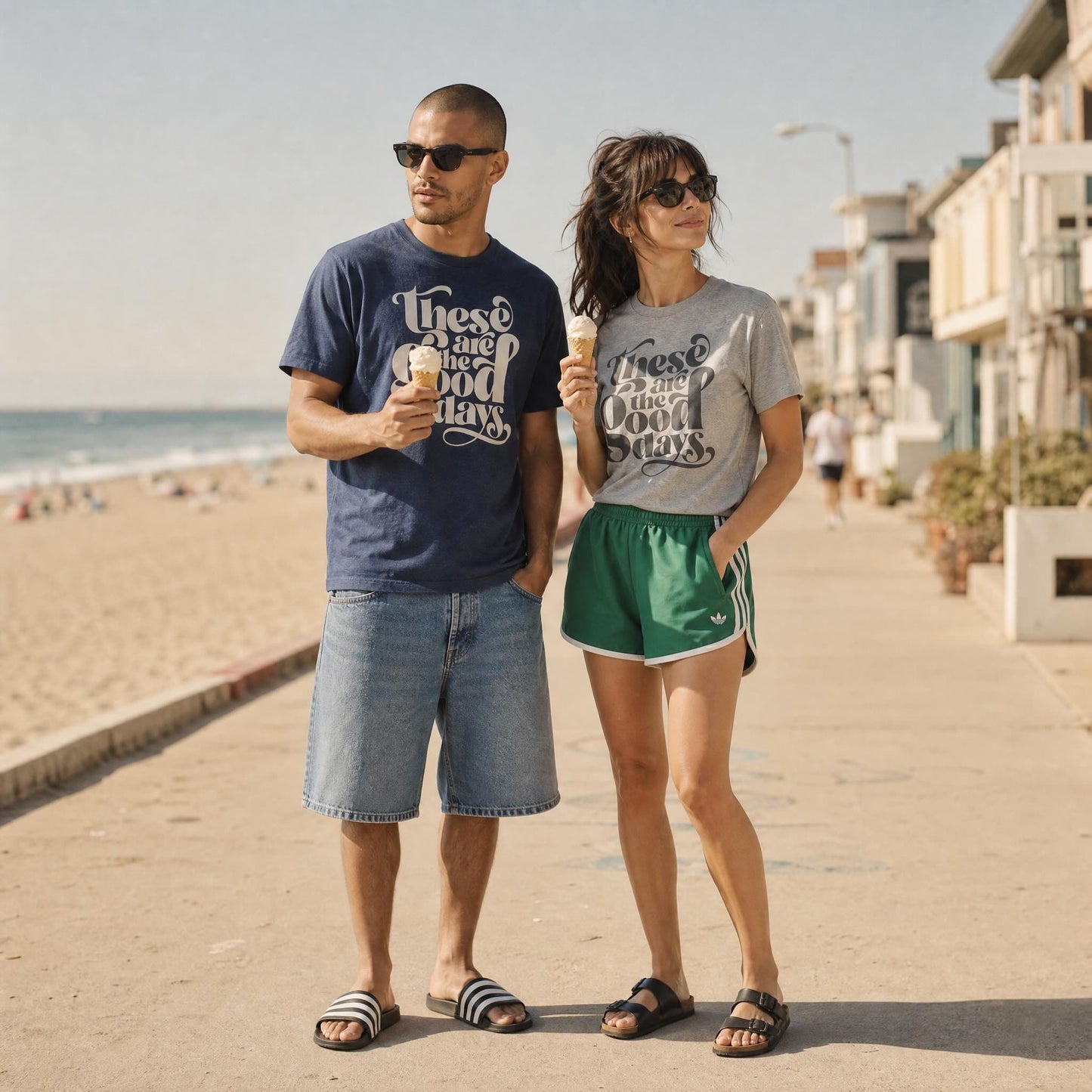Couple wearing These Are The Good Days graphic t-shirts on a sunny beachside promenade, eating ice cream. Man in navy marl  tee and denim shorts, woman in grey marl tee with green Adidas shorts and Birkenstock sandals.