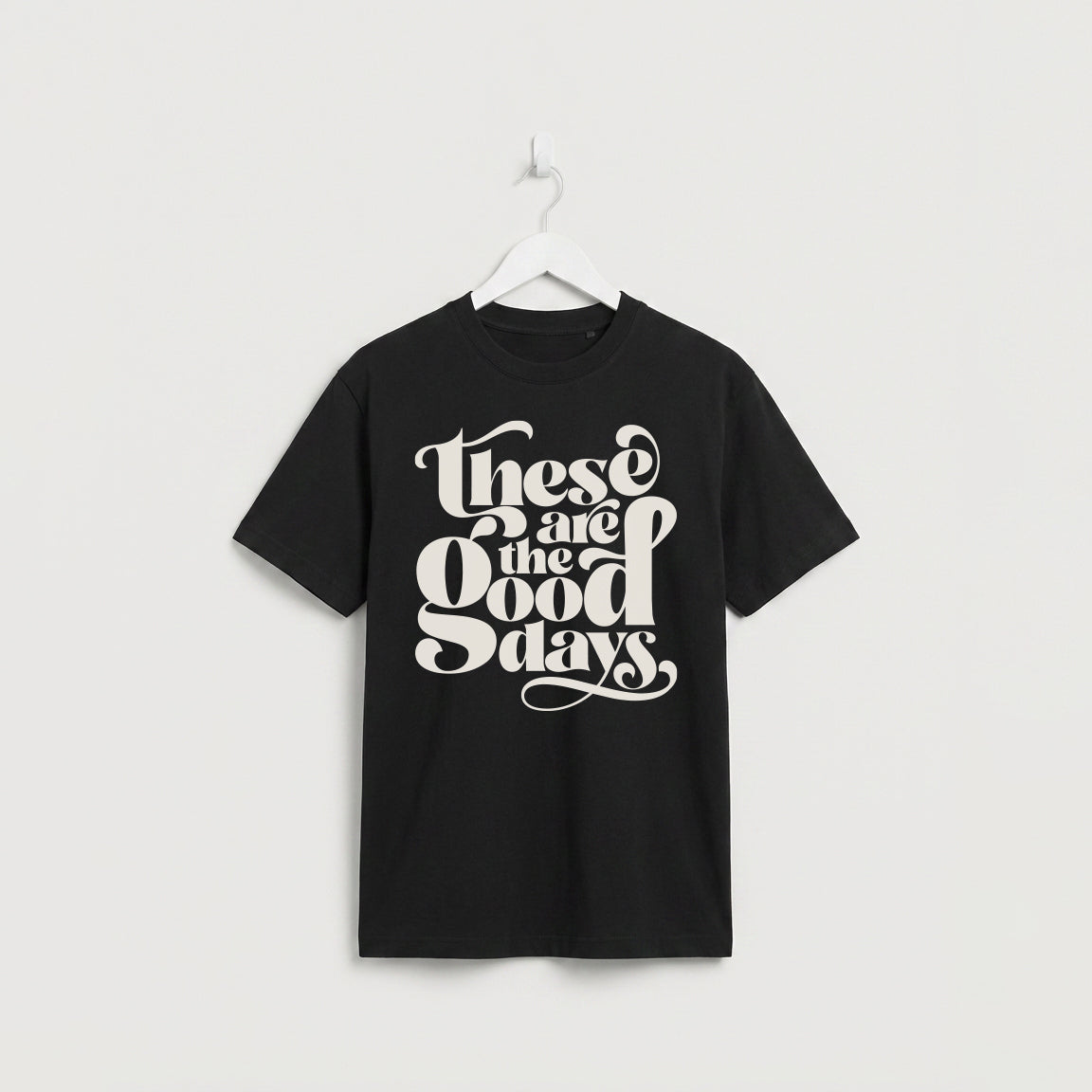 Black “These Are the Good Days” graphic t-shirt on white wooden hanger, minimalist studio background