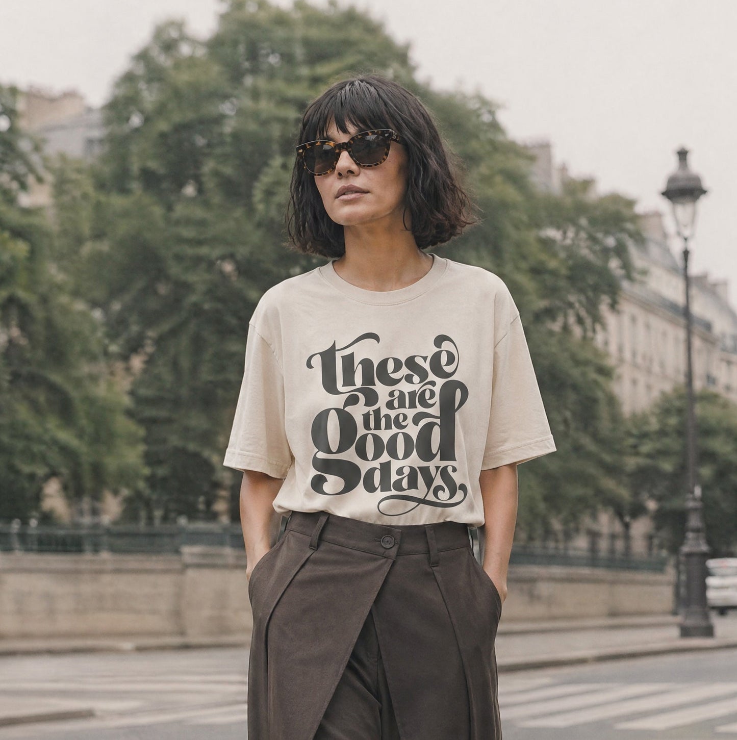 Model with brown bob hair cut, wearing ecru coloured graphic  t-shirt with 'These are the good days" quote in bold calligraphy typography, tucked into brown barel leg trousers. 
