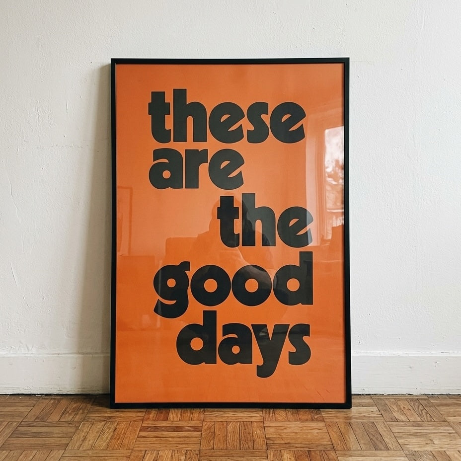 Orange and black “These Are The Good Days” typography art print in a slim black frame, leaning against a white wall on a parquet wooden floor.