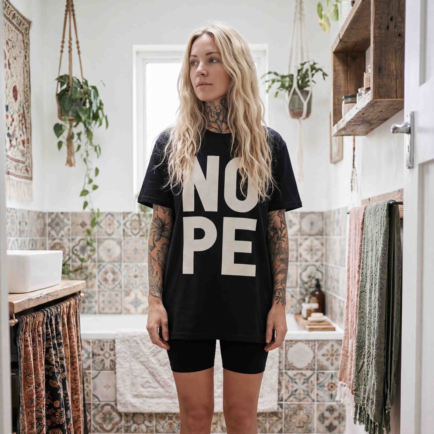 Blode girl with nature tattoos standing in a bohemian styled bathroom, wearing ablack NOPE tshirt with bold type, and black cycling short. 