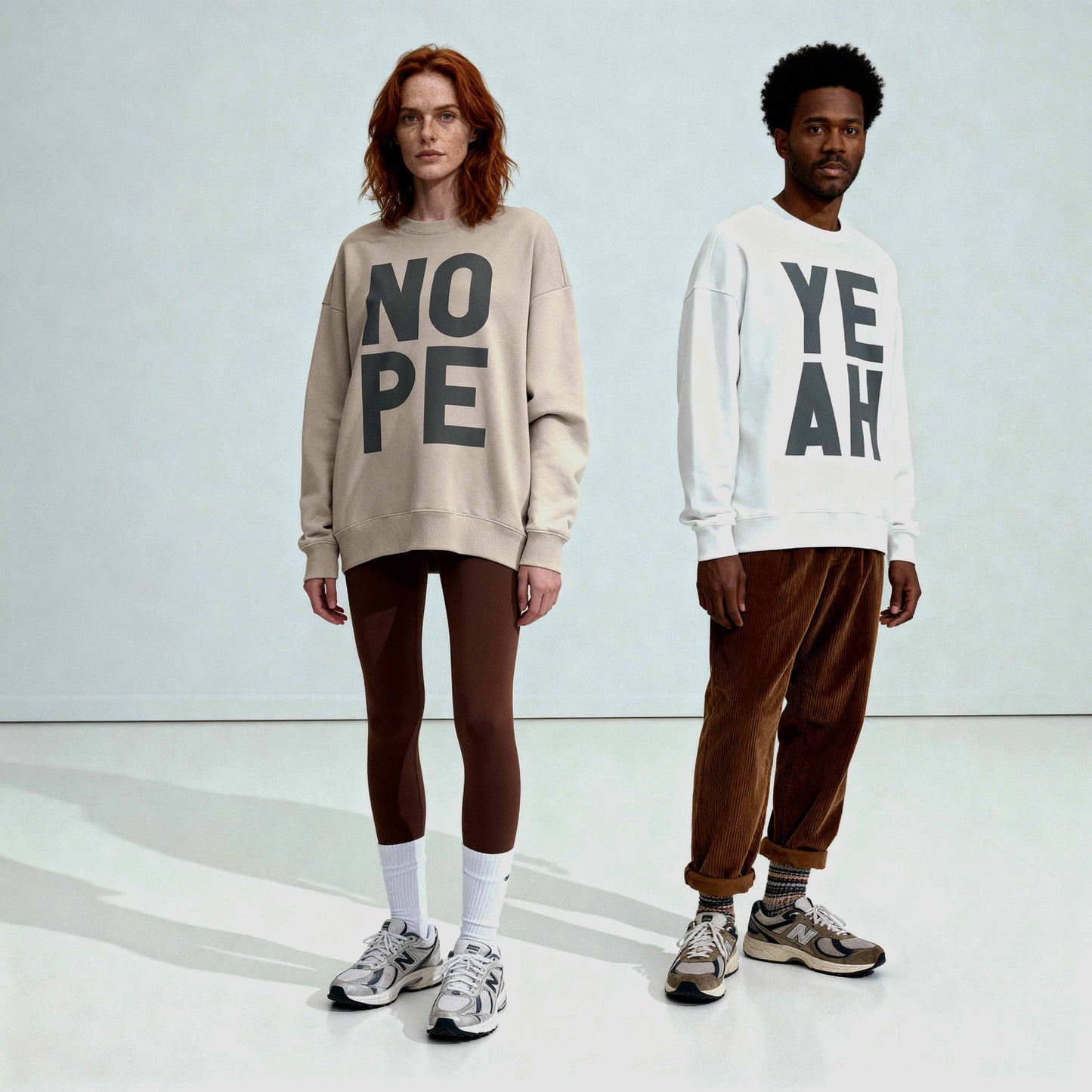 Auburn haired model with freckles wearing Beige colour oversized NOPE sweatshirt with leggings and sports socks, next to black man wearing white YEAH sweatshirt with cords and trainers. 